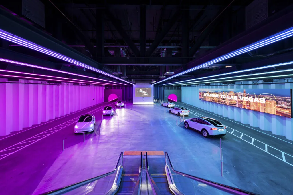 Center Loop Stop at the Las Vegas Convention Center. The interior is illuminated in hues of pink and purple, and Tesla vehicles await passengers with two tunnel openings in the background.