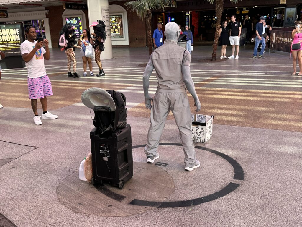 A street perormer dressed in gray and with skin painted in gray poses, waiting for a tip to start the show.