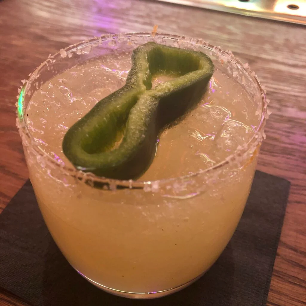 A yellow cocktail, garnished with a slice of green pepper on top. 