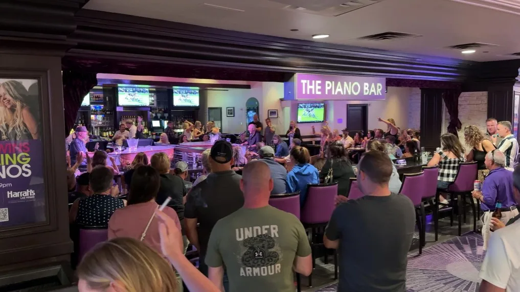 A crowd gathers around the piano bar. Inside are two pianos being played by performers.