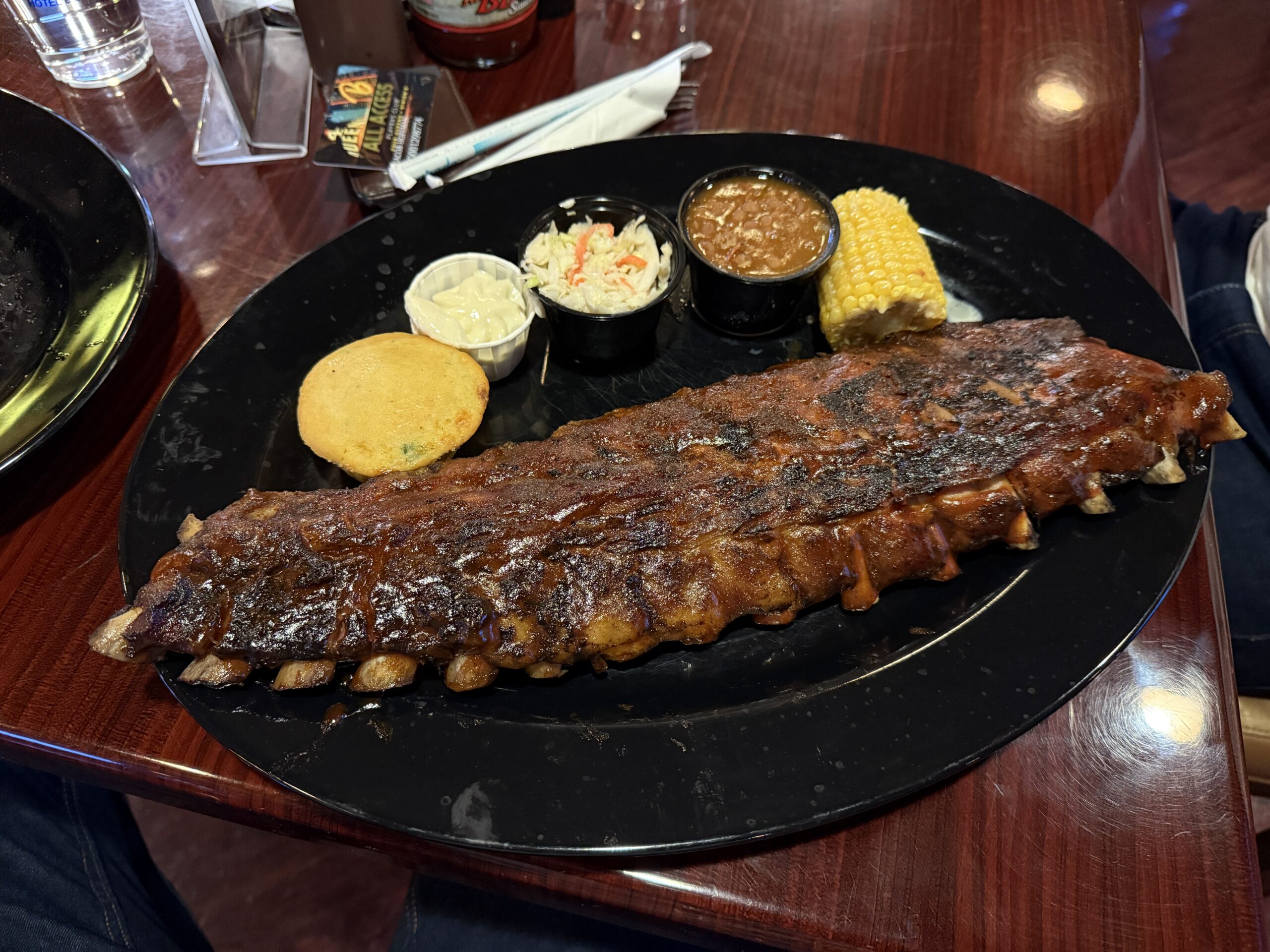 A rack of ribs, corn on the cob, and corn bread on a black plate. 