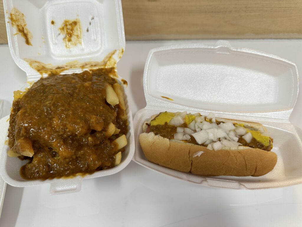 A styrofoam container of fries covered in chili next to a chili dog. 