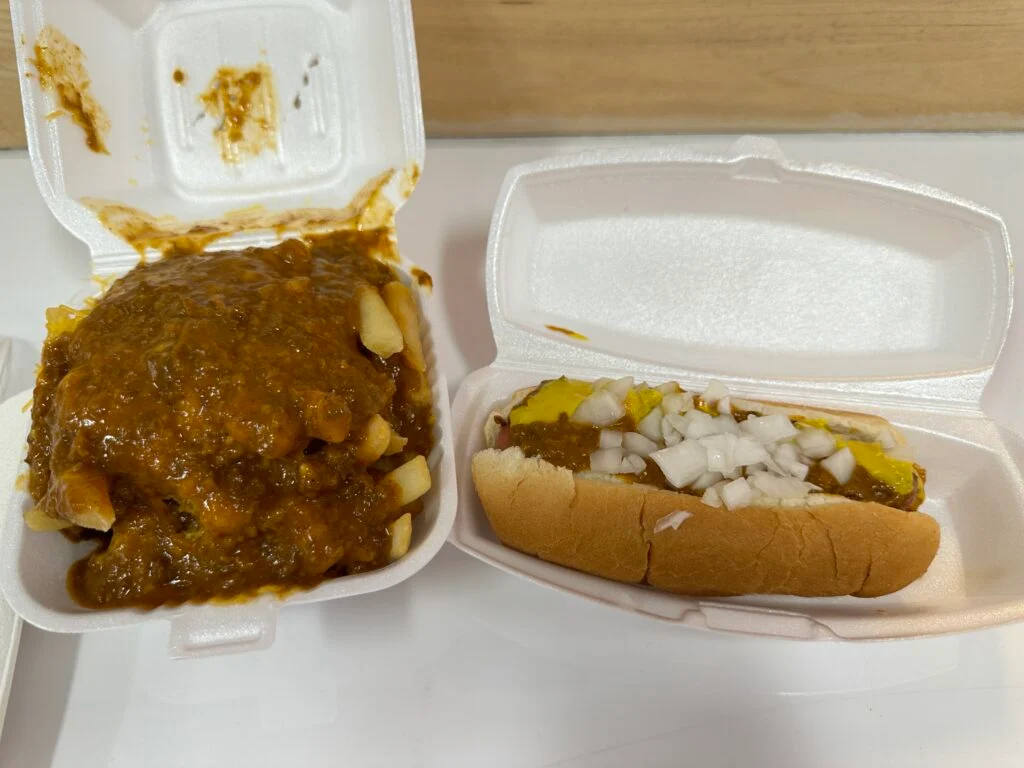 A styrofoam container of fries covered in chili next to a chili dog. 