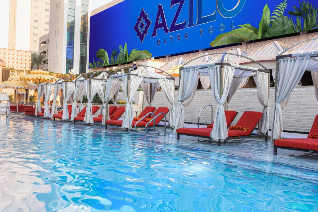 White cabanas with red seating surround the blue water of Azilo Pool. Above the cabanas is a long TV screen.