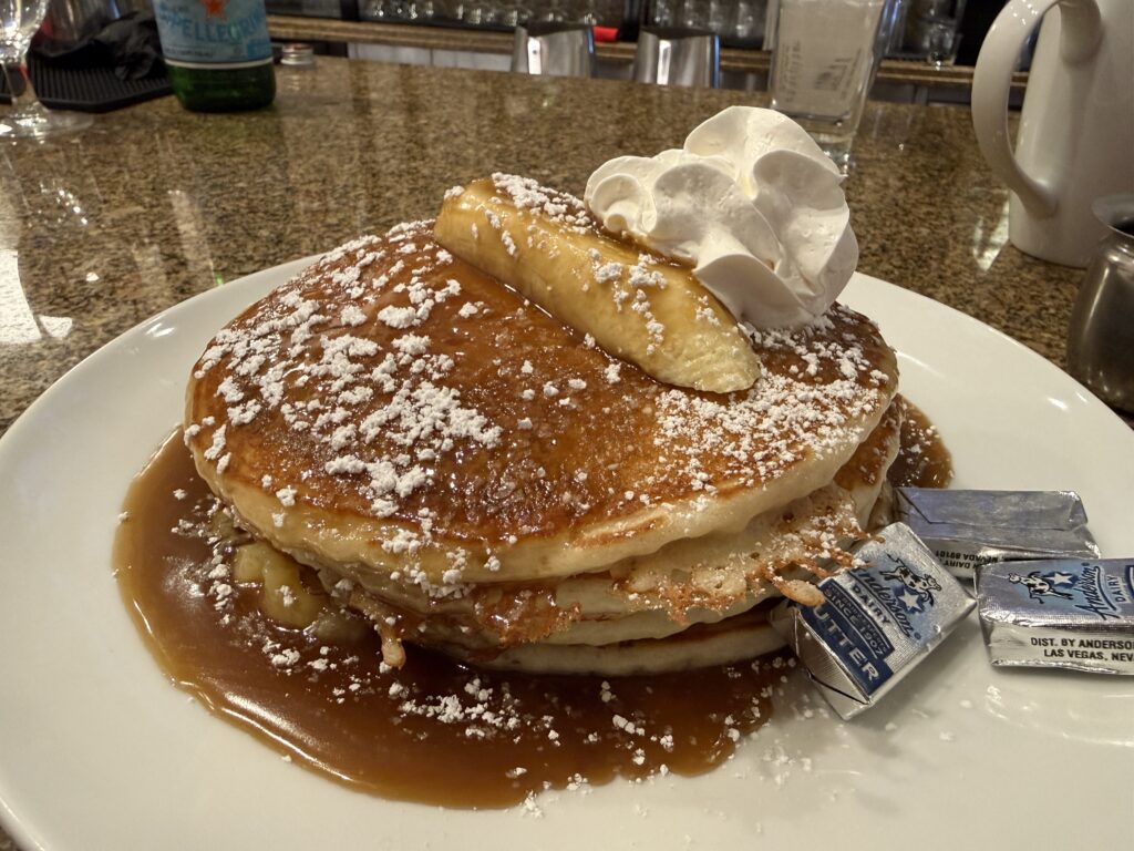 A stack of 3 pancakes topped with a banana, whipped creme, caramel, and powdered sugar.