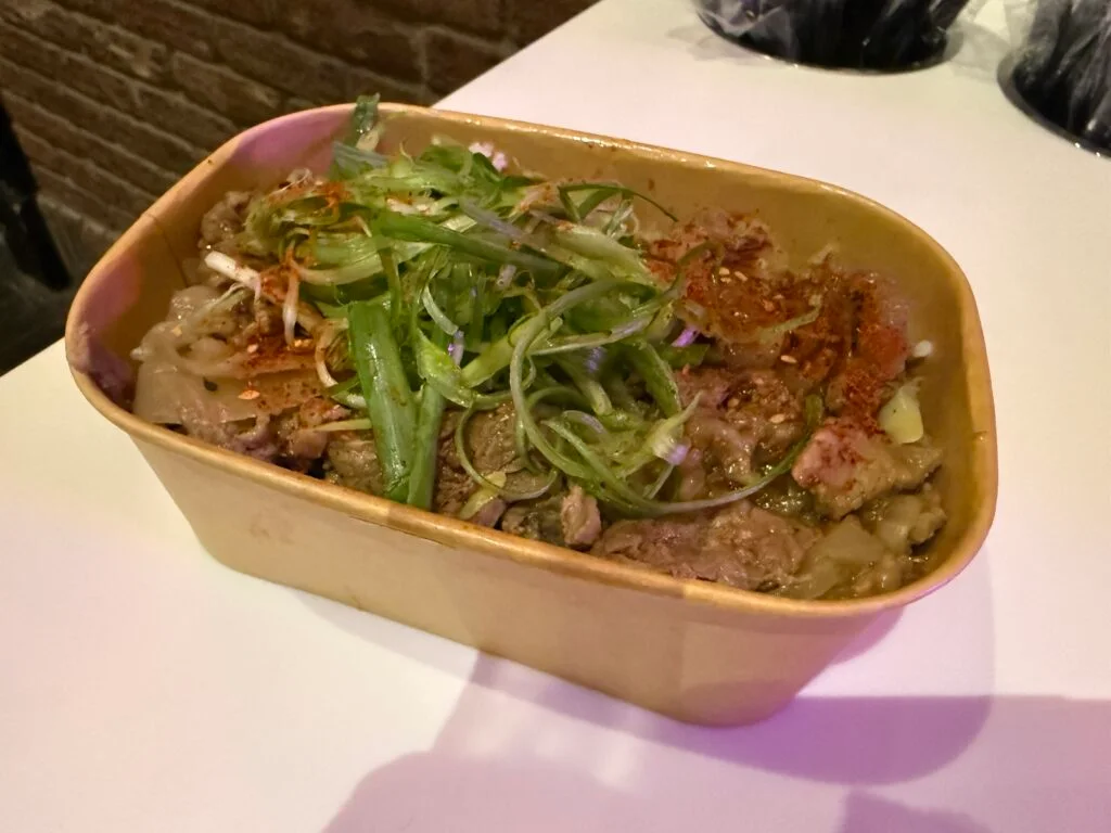 A square shaped bowl with beef, rice, and pickled ginger.