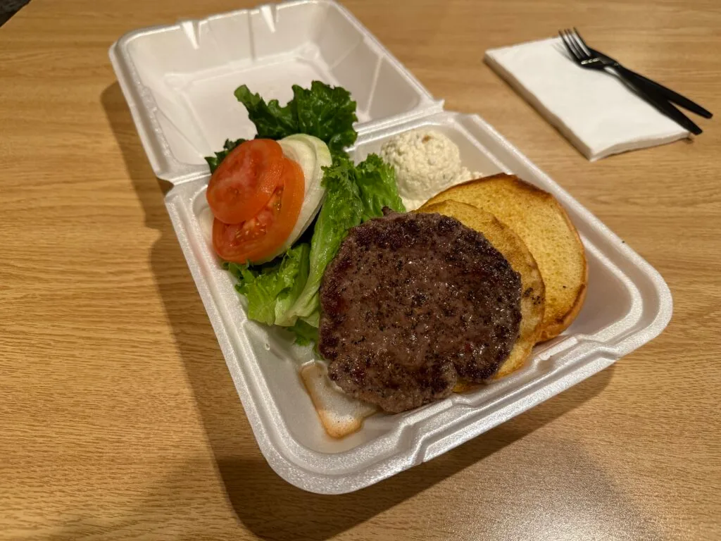 An open faced burger with a stack of veggies to the side of it alongside a little mound of potato salad. 