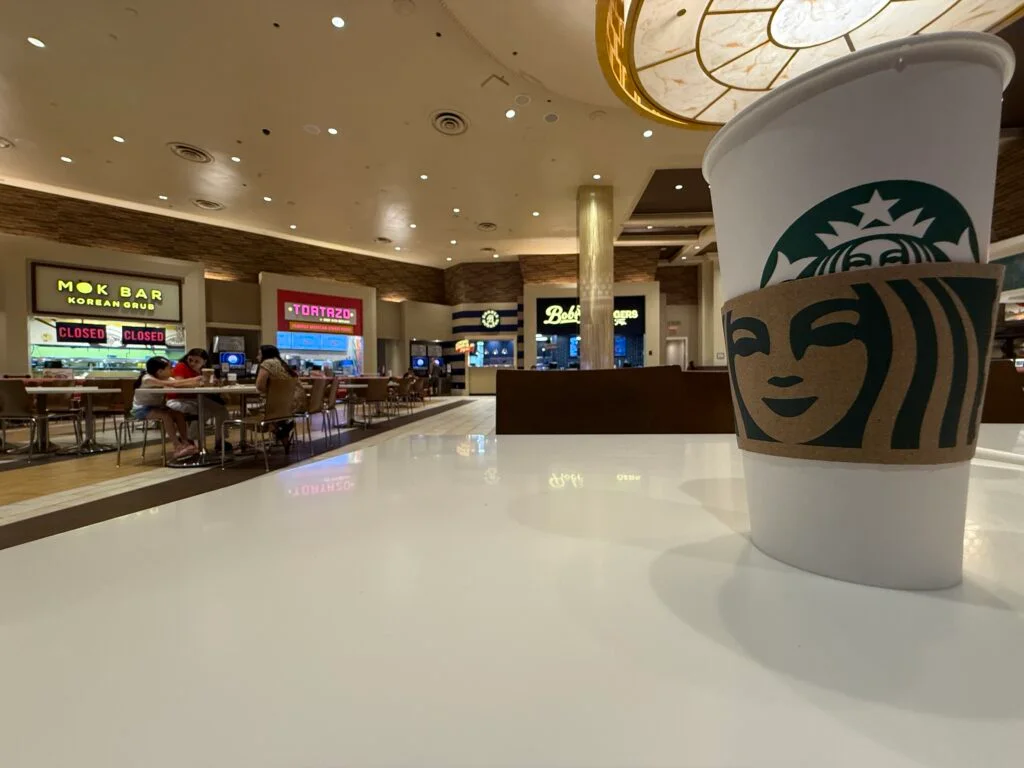 A cup of coffee on a table, behind it are the food outlets in the court.