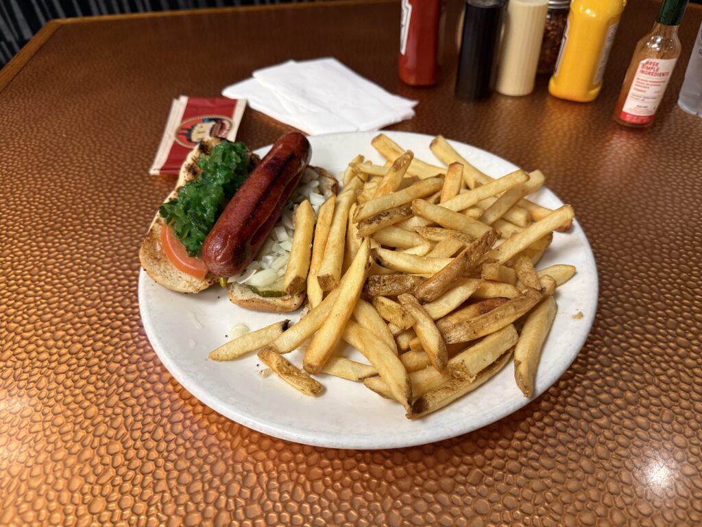 A chicago dog on a plate next to a large mountain of french fries. 
