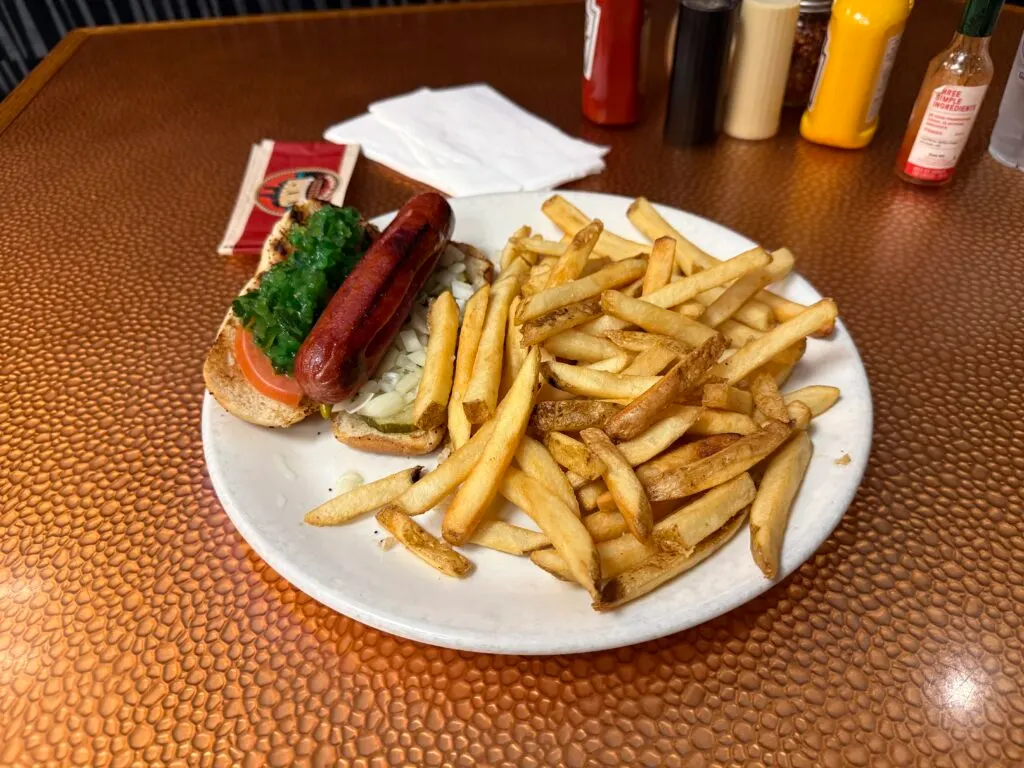 A chicago dog on a plate next to a large mountain of french fries. 