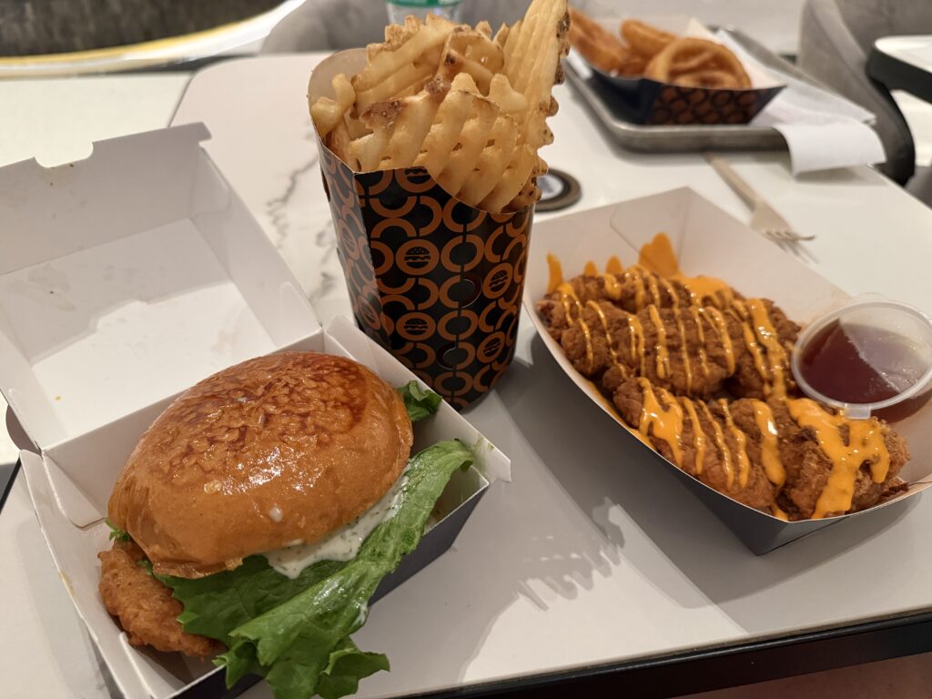 A chicken sandwich in a box, a cone full of waffle fries, and a basket of 3 chicken tenders with a buffalo drizzle. 