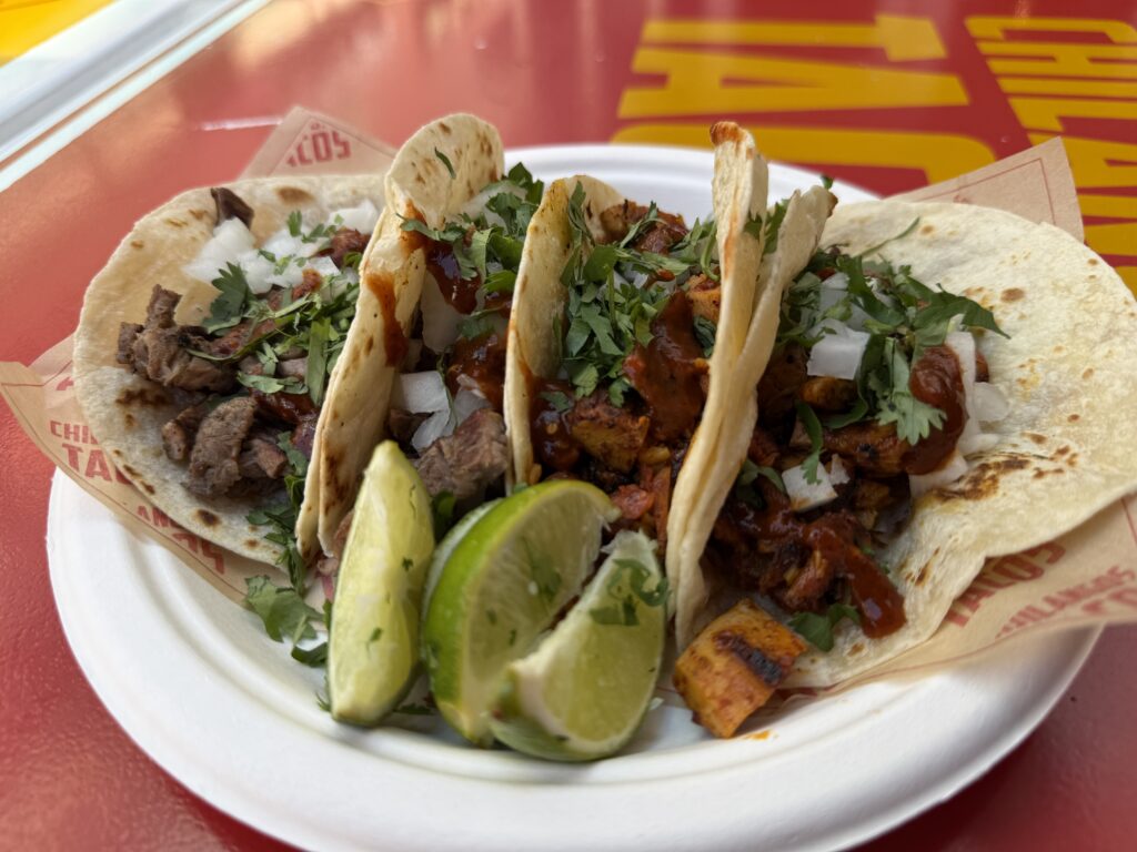 Soft tacos filled with chopped meat, onions, salsa, and cilantro on a red table with lime wedges nearby