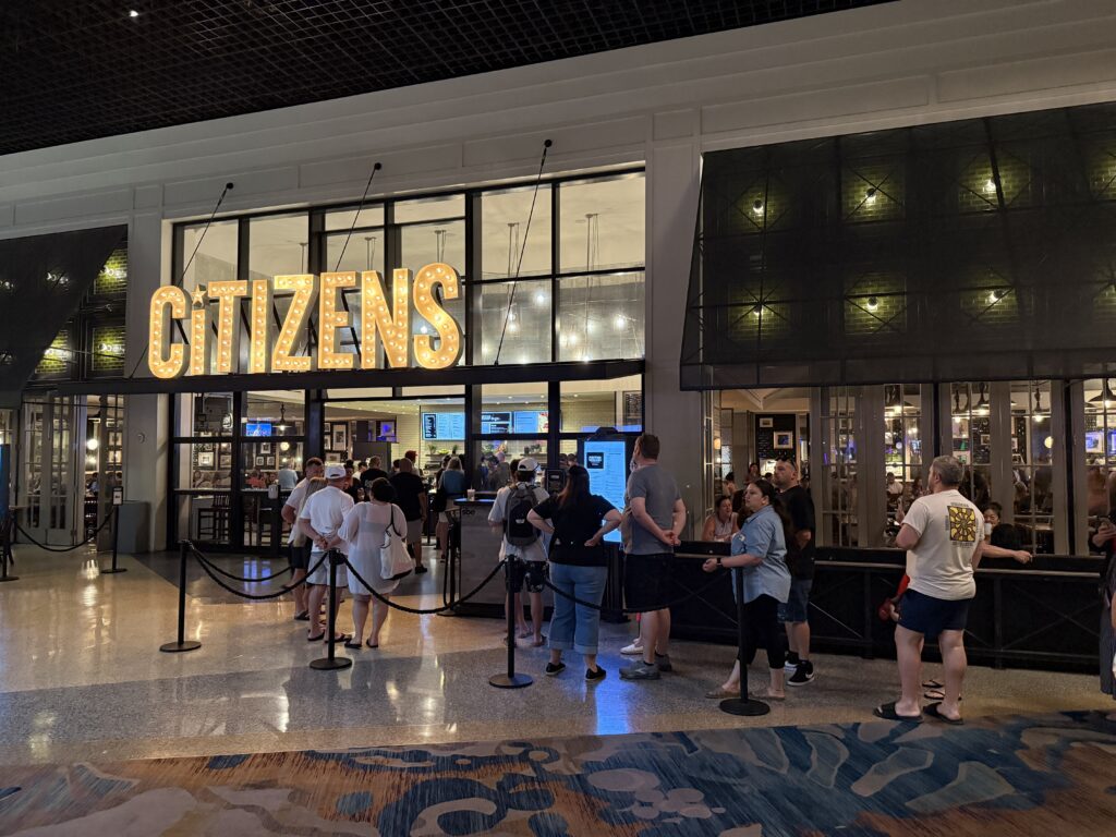 People waiting in a lined queue inside a modern building with a large illuminated 'CITIZENS' sign visible above the counter area