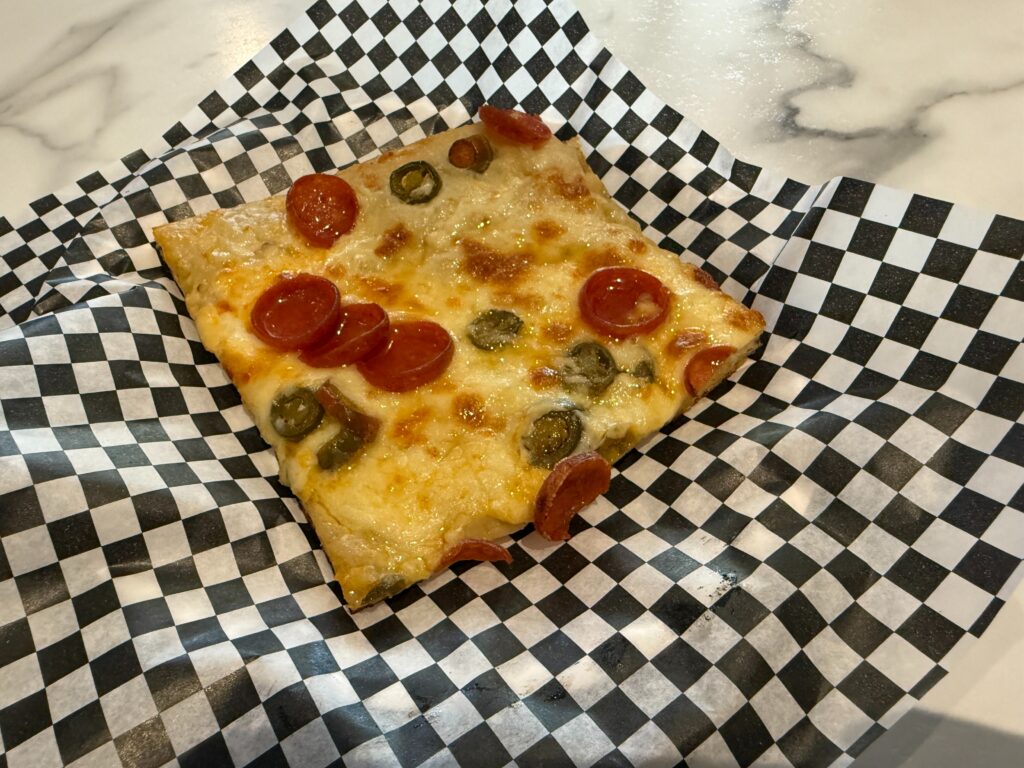 A square slice of pizza on a checkered piece of paper topped with pepperoni and jalapenos. 