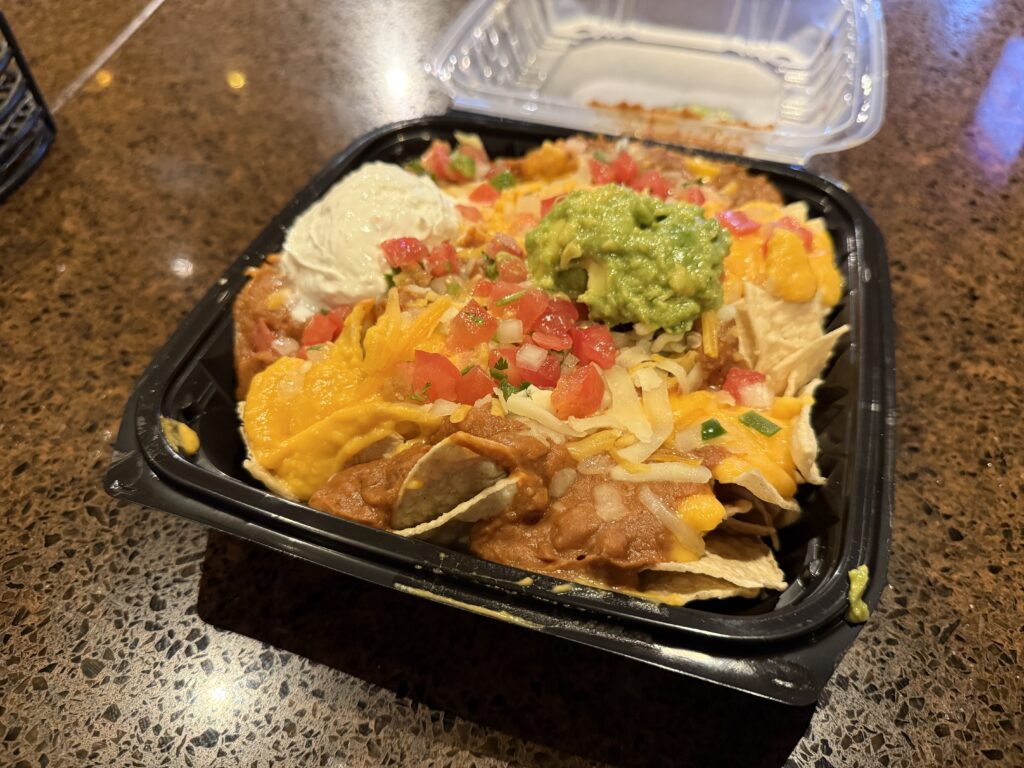 Nachos in a black plastic tray, topped iwth cheese, tomatos, guac, and sour cream. 