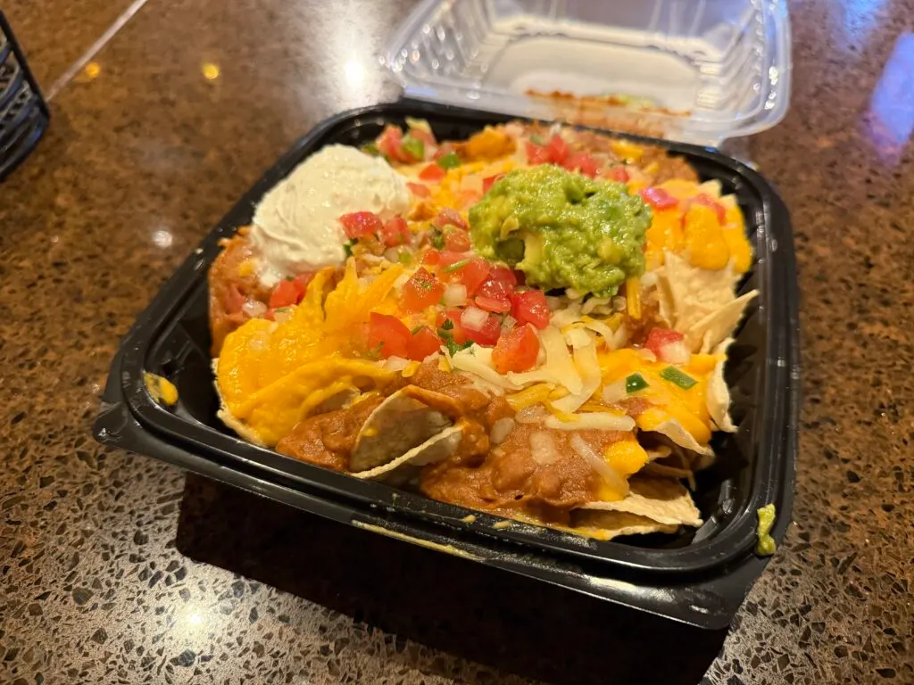 Nachos in a black plastic tray, topped iwth cheese, tomatos, guac, and sour cream. 