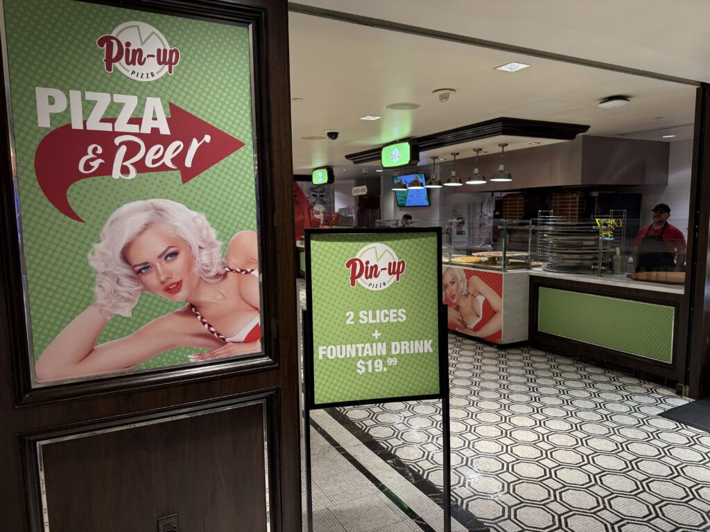 A sign in front of Pin-Up Pizza advertising a 2 slices and a drink deal.
