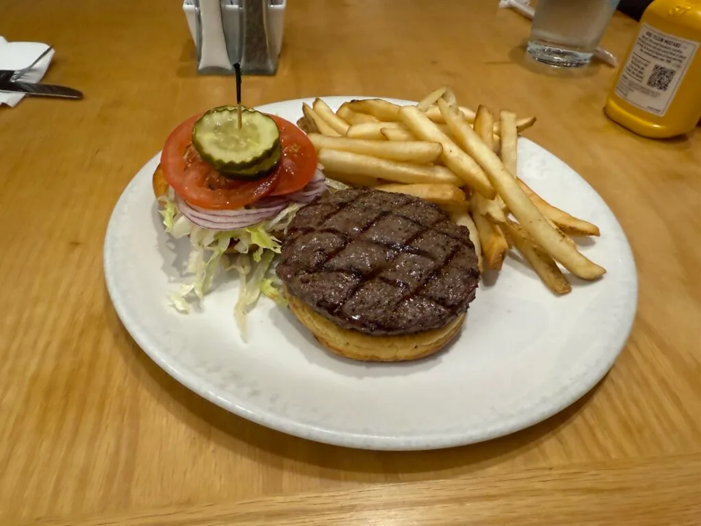 An open faced burger next to the top of it's bun, which is stacked with lettuce, tomato, and pickles next to a pile of french fries. 