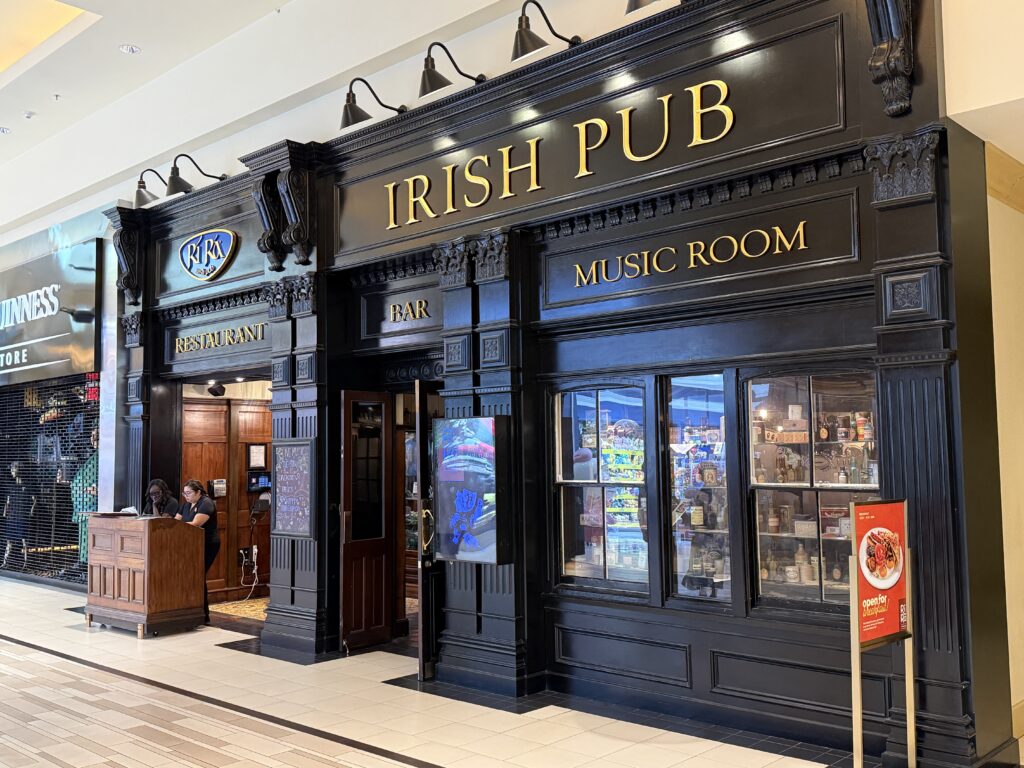 Ornate black pub storefront with gold IRISH PUB and MUSIC ROOM signs, glass display windows, and two staff at a wooden reception desk inside a mall.