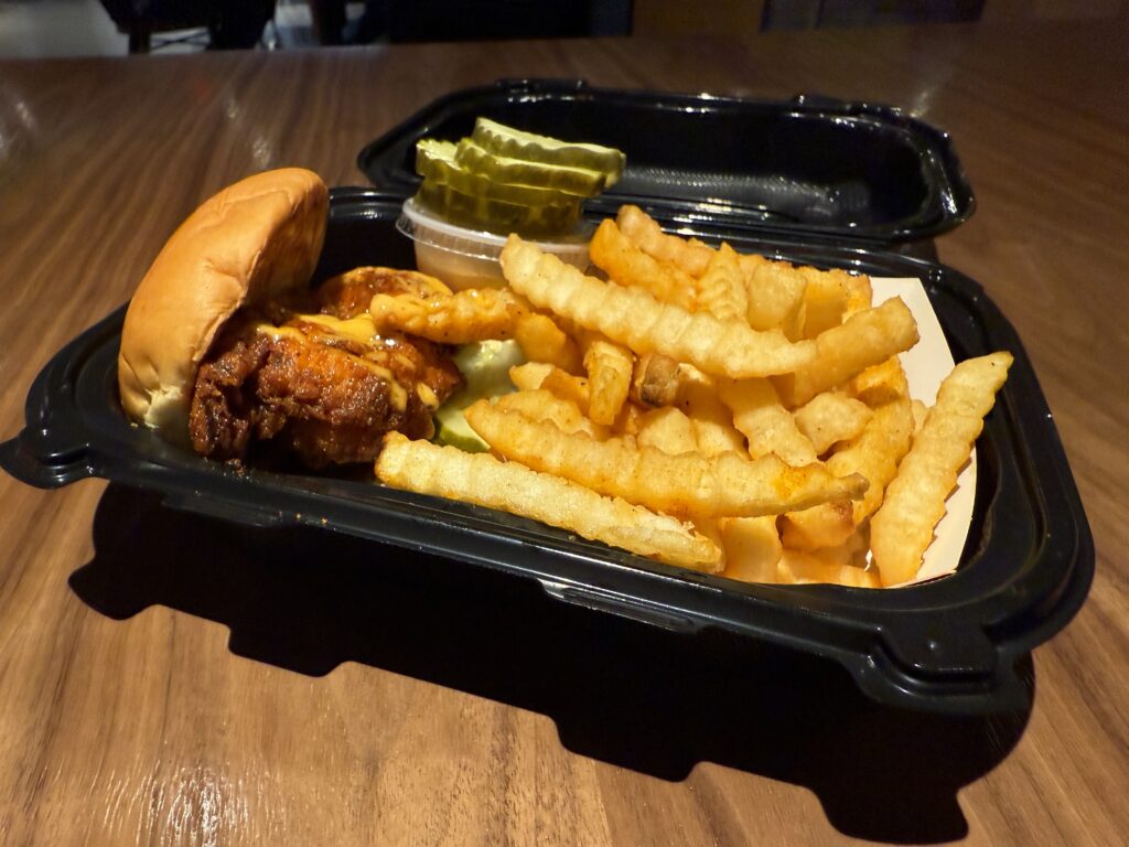 A chicken slider next to fries and a stack of pickle chips in a black tray. 