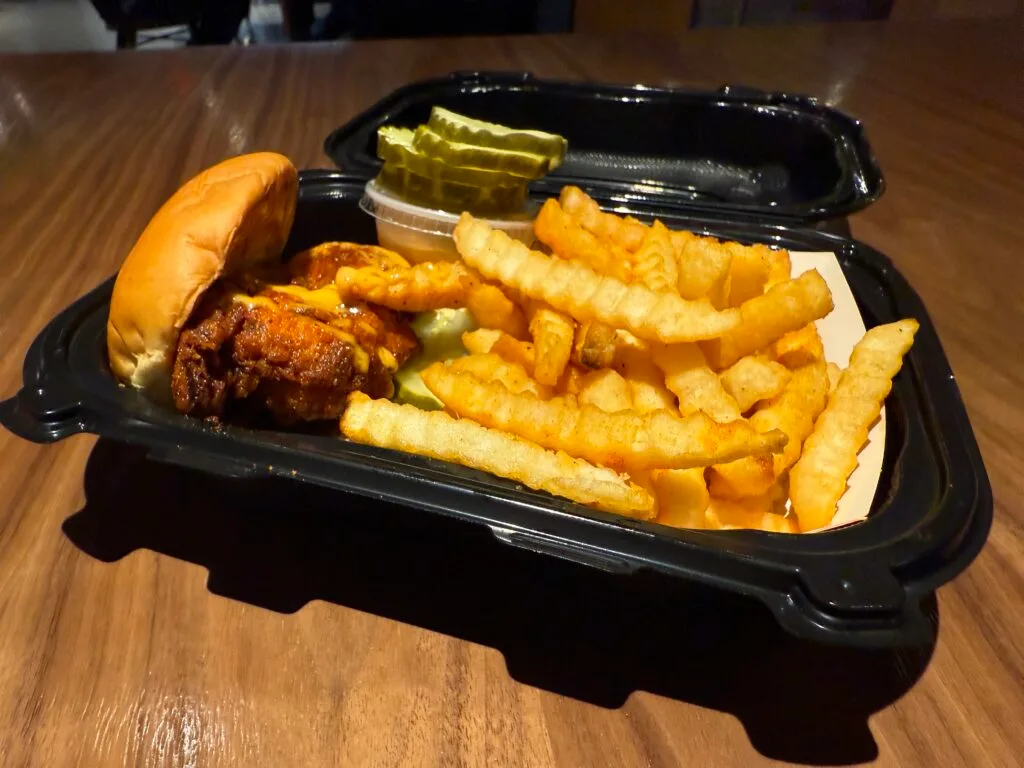 A chicken slider next to fries and a stack of pickle chips in a black tray.