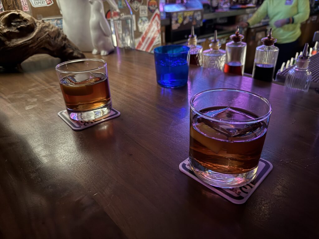2 old fashioneds sit site by side, each with a massive square ice cube in the glass.