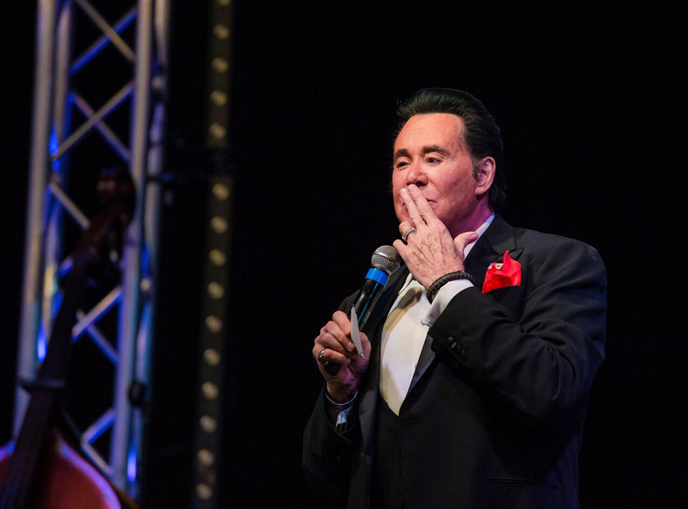 Wayne newton puts his fingers to his mouth on stage