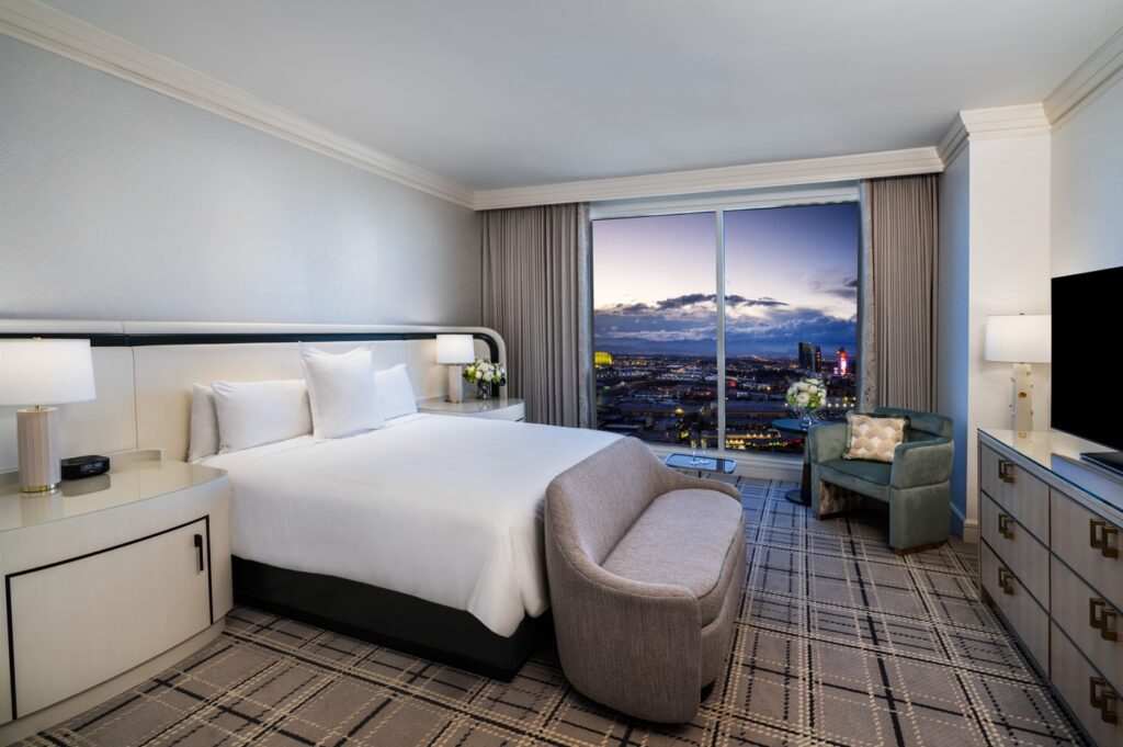 A king bed with a white headboard. An open window in the backgrownd offers a view of the Strip.