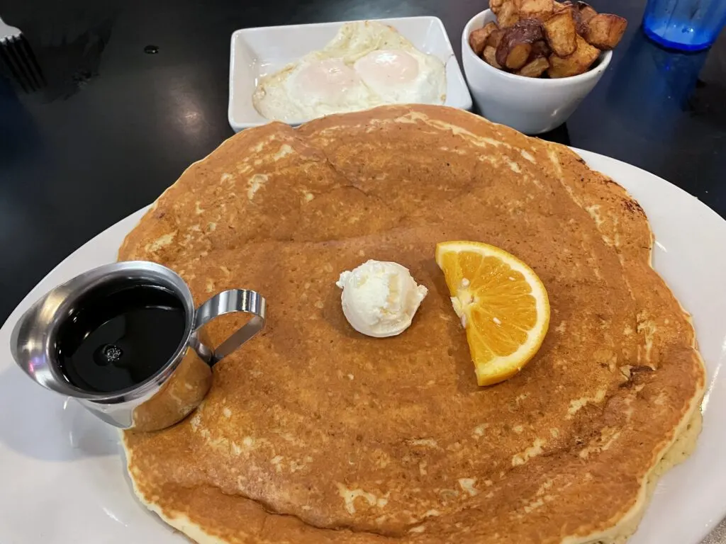 A large pancake, 2 eggs over easy, cubed potatoes, syrup, and an orange slice on a plate