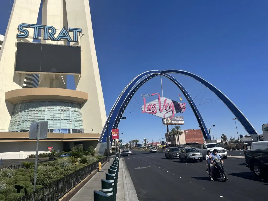 A decorative gateway arch stands over Las Vegas blvd. with the base of STRAT's tower to the left.