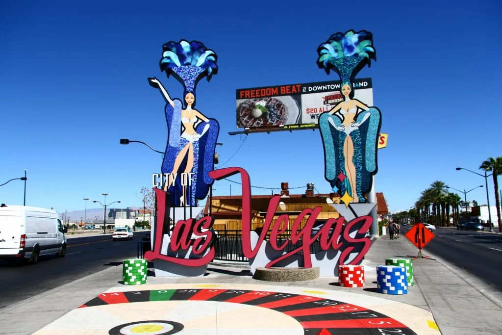 A sign that says "City of Las Vegas" with 2 large showgirls behind it and a roulette wheel painted on the ground in front. 