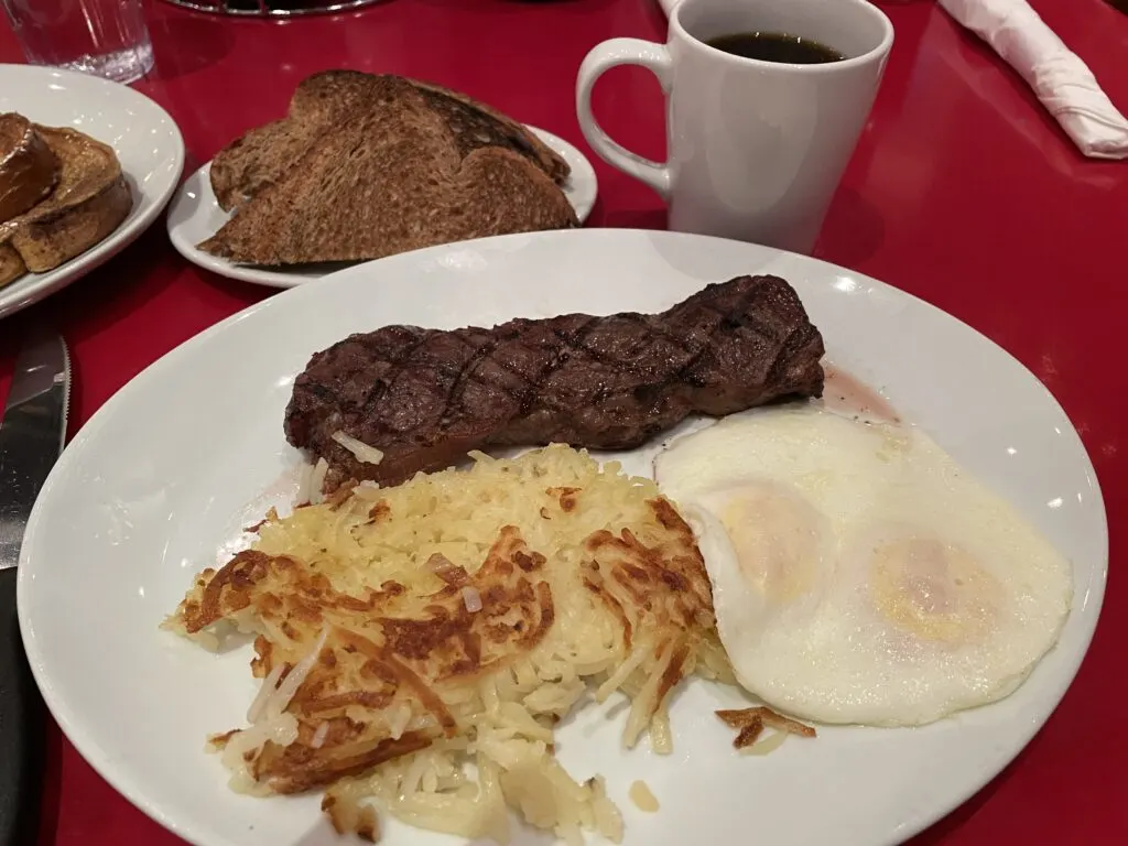 A steak, pile of hashbrowns, and 2 eggs over easy on a plate with toast and a coffee mug to the side.