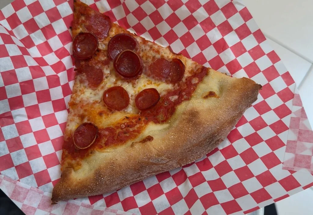 A pepperoni slice of pizza on a checkered paper on a plate. 