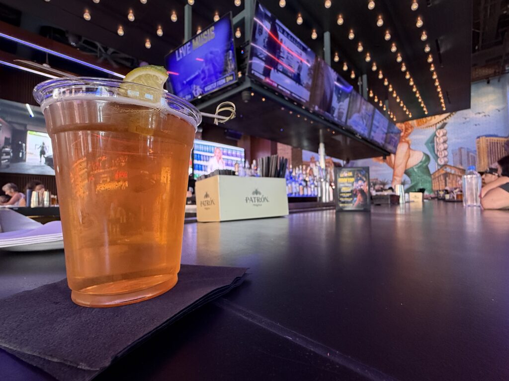 A beer, on the bar at bottled blonde with TVs behind it playing sports.