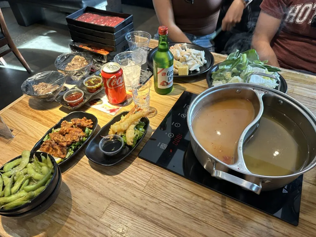 A kettle full of broth stands ready on a burner, surrounded by veggies and meat to be consumed.