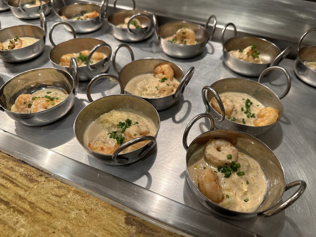 A number of metal bowls holding shrimp & grits, which are drenched in a white sauce.