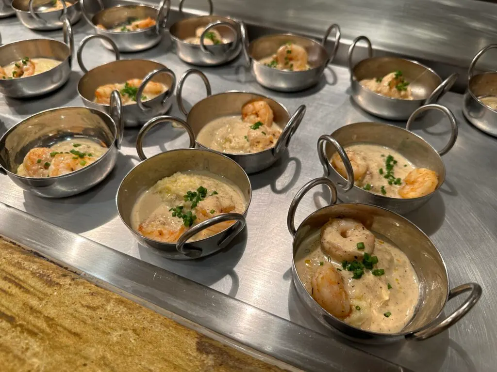 A number of metal bowls holding shrimp & grits, which are drenched in a white sauce.