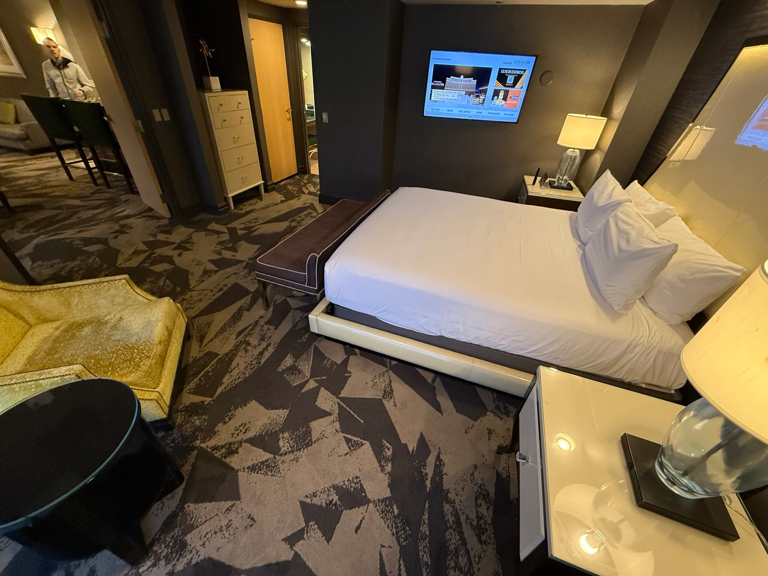 A king bed with a TV mounted on the wall to the side of it. Doorways to the bathroom and living area can both be seen. 