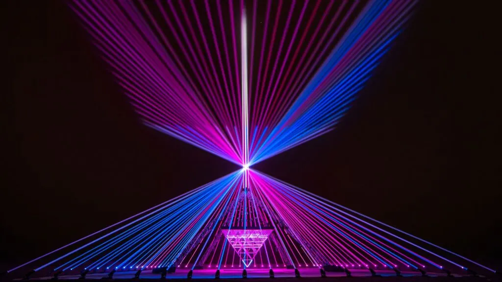 A large piece of art which is comprised of purple and blue laser lights shooting up and intersecting eachother in a triangle formation. Inside the illuminated triangle is an upside down triangle balancing on its point constructed of metal bars.