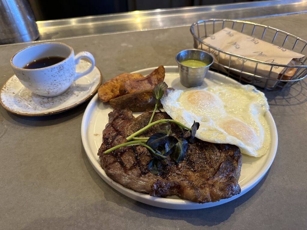 A large ribeye steak sharing a plate with two over easy eggs and potatoes.