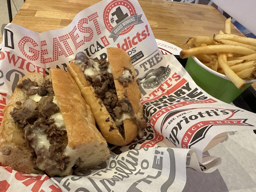 A philly cheesesteak on a tray next to a cup of french fries.