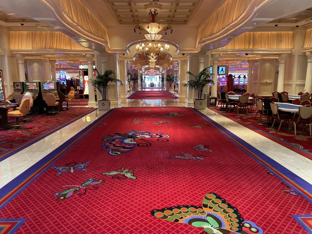 Gaming floor at Encore. On the floor is red carpet with butterflies and marble. Slot machines line the left side and poker tables are on the right.