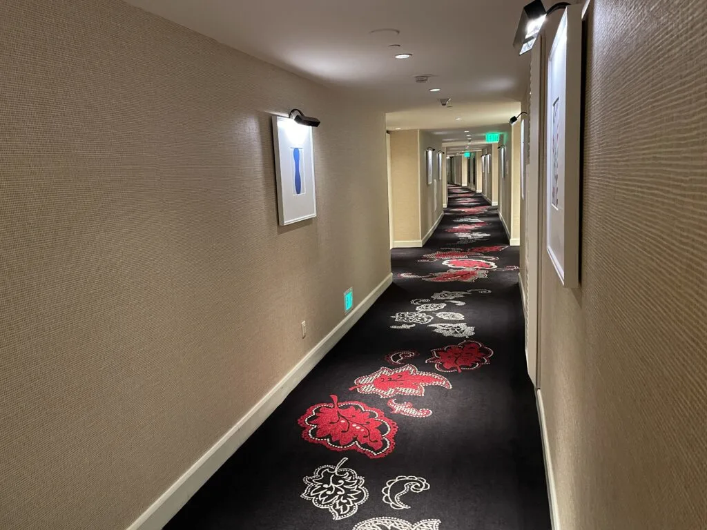 AHotel hallway with black carpet accented by a  white and red flower pattern. The walls are lined with a series of small paintings illuminated by a light above each.