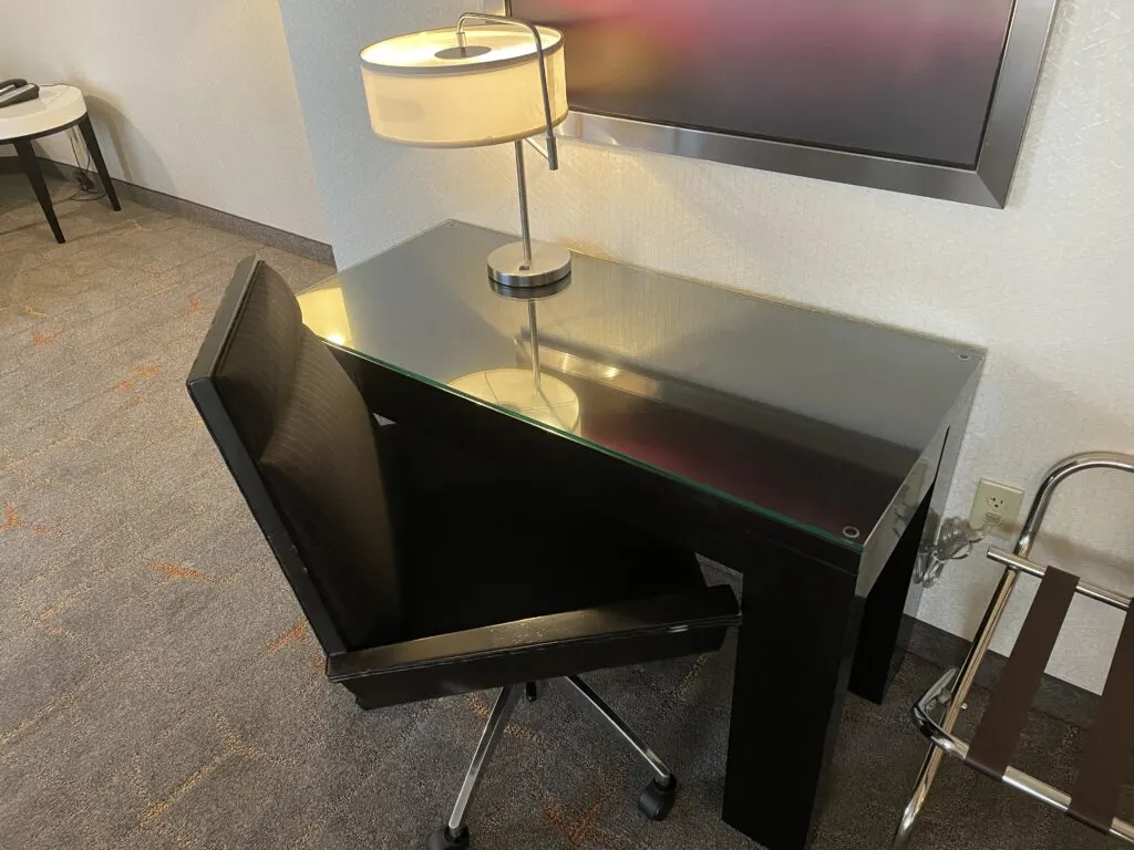 A dark, glass topped workdesk and chair. On the workdesk is a lamp.