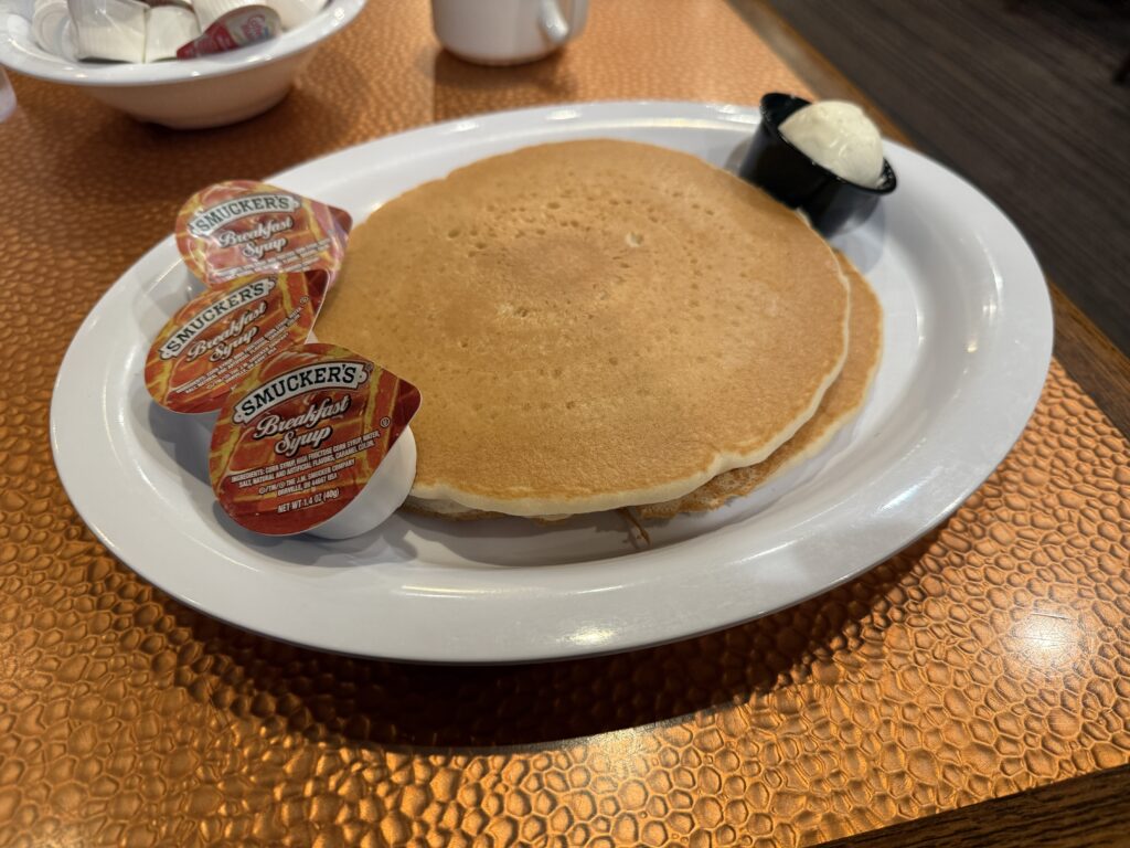 A stack of pancakes on a white plate with a container of butter and 3 plastic containers of syrup.