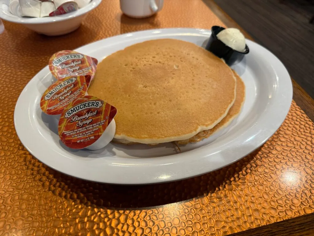 A stack of pancakes on a white plate with a container of butter and 3 plastic containers of syrup. 