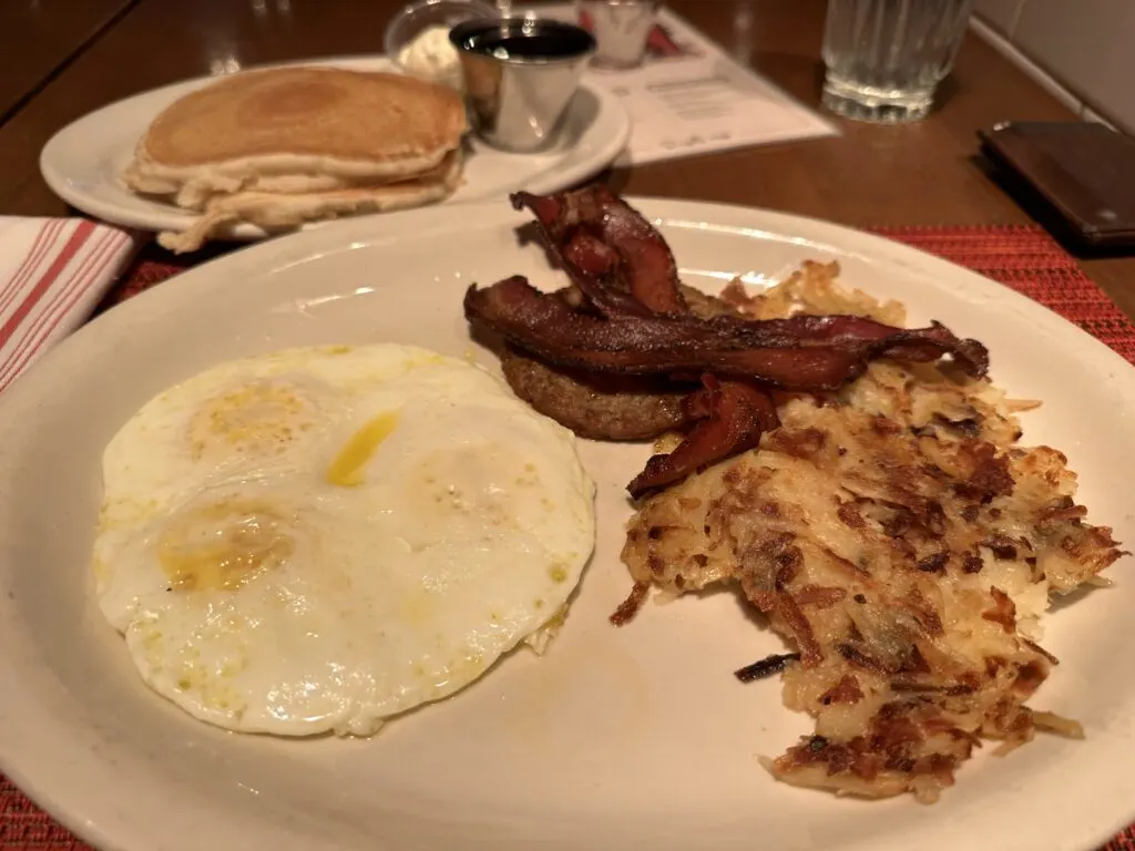 3 over easy eggs, bacon, and hashbrouns are on a plate. Behind it is a plate with 2 pancakes and a cup of coffee.