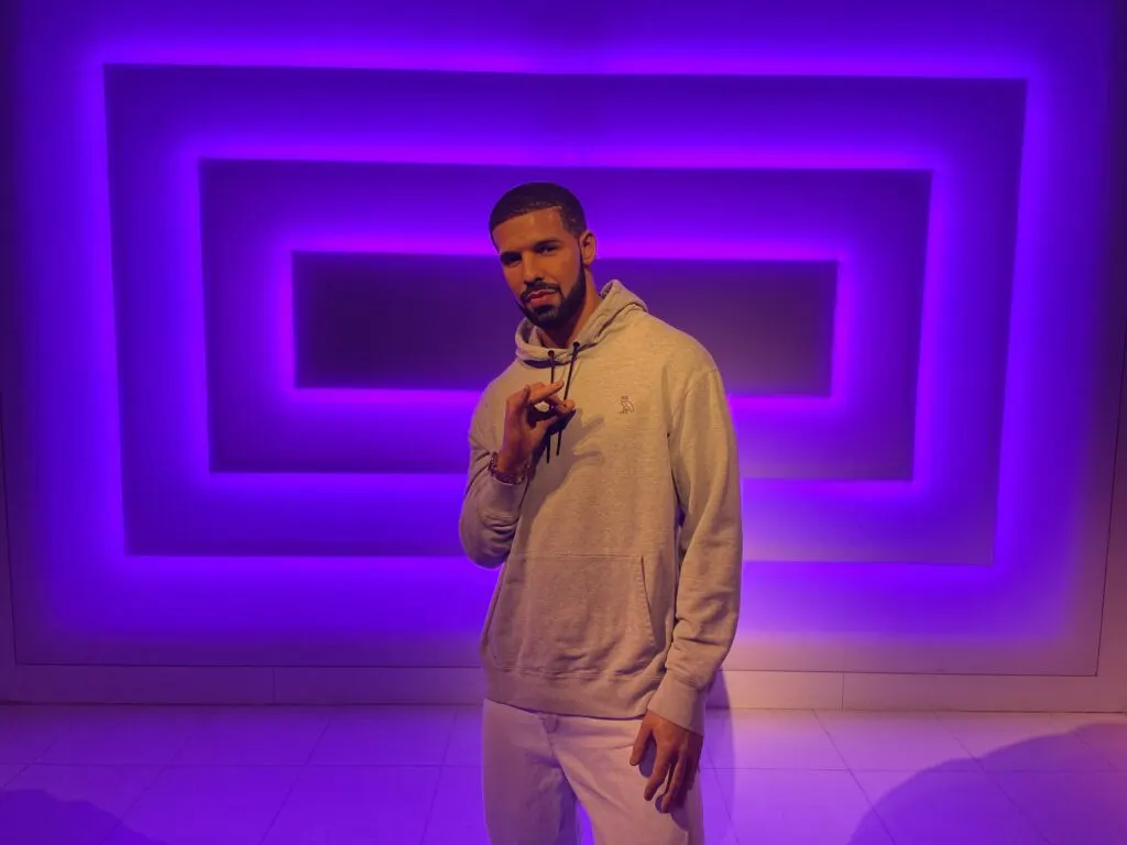 Drake poses in front of a purple illuminated backdrop wearing a light colored sweat suit.