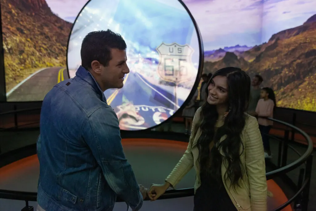 Two people watching a circular TV screen with a larger one behind it on the wall