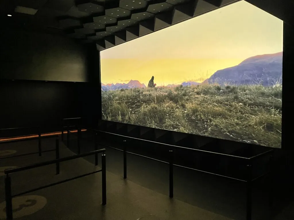 An empty room with a large theater style screen depicting a man sitting at a fire in the mountains. 